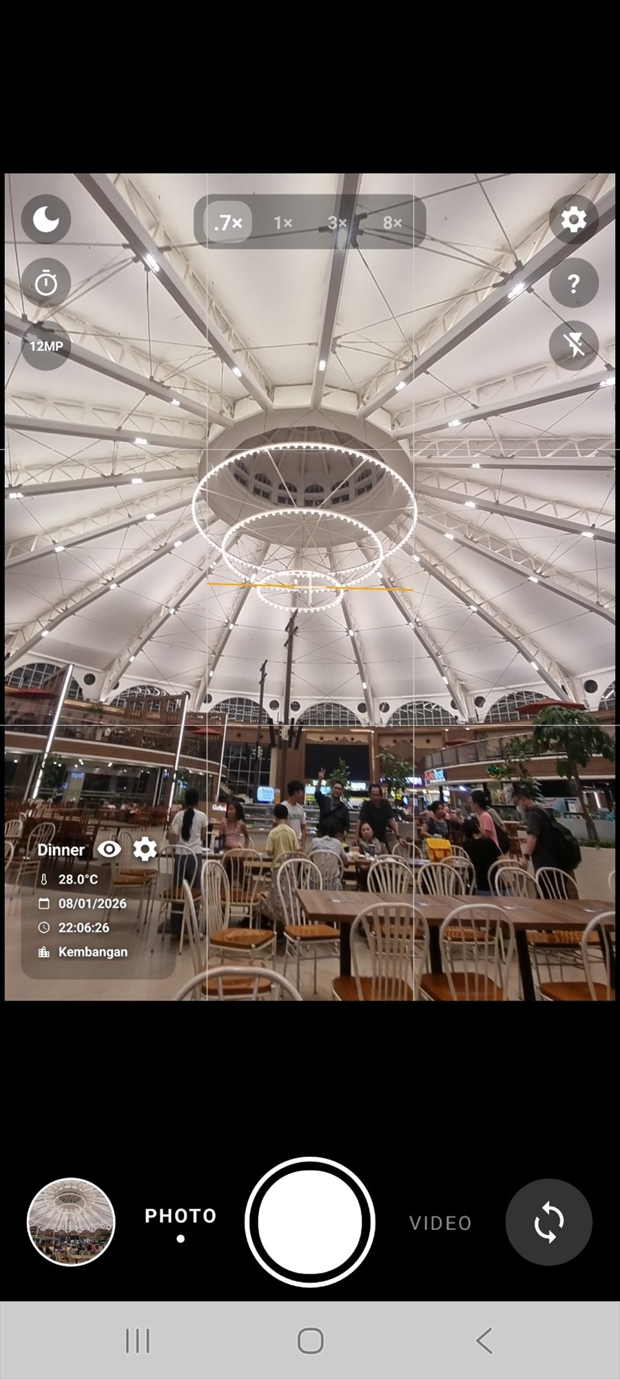 An indoor food court photographed in the evening, stamped with the event name Dinner, 28.0 degrees Celsius, the date, the time and the city Kembangan.
